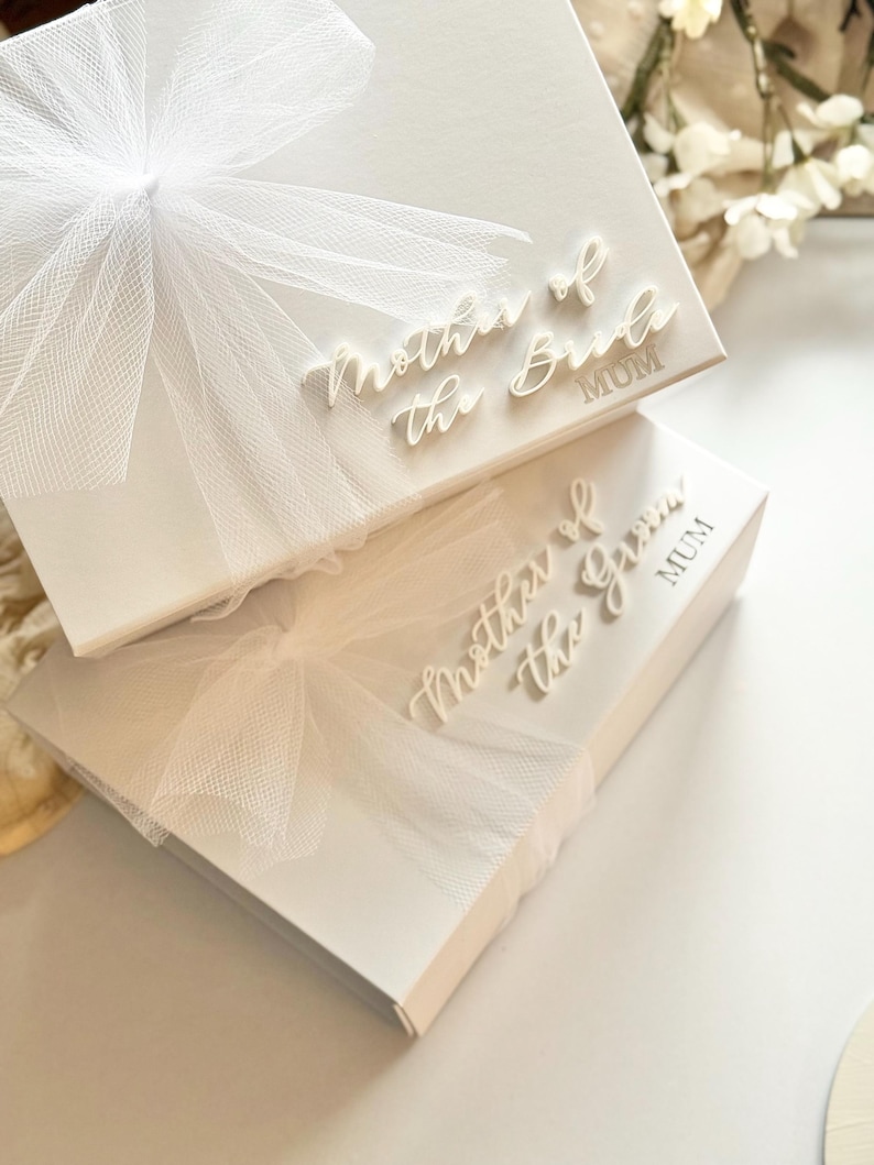 K&ouml;nnte beinhalten: Zwei wei&szlig;e Geschenkboxen mit den Schriftz&uuml;gen "Mother of the Bride" und "Mother of the Groom" in Schreibschrift. Jede Box ist mit einer wei&szlig;en T&uuml;llschleife verziert. Das Wort "MUM" ist darunter gedruckt.