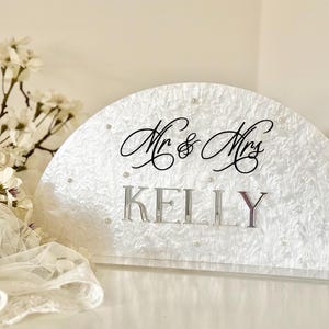 May include: A semi-circular wedding sign with the words "Mr & Mrs" in elegant script above the name "KELLY" in silver block letters. The sign has a textured white surface and is adorned with small pearls.
