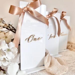 Könnte beinhalten: Drei weiße Tischnummernschilder mit goldenen Kalligraphie-Zahlen. Die Zahlen sind "Eins", "Zwei" und "Drei". Jedes Schild hat ein beigefarbenes Satinband, das zu einer Schleife gebunden ist.