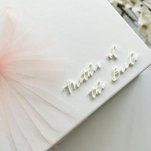 K&ouml;nnte beinhalten: Eine wei&szlig;e Geschenkbox mit einer zarten rosa T&uuml;llschleife. Die Worte &bdquo;Mother of the Bride&ldquo; sind in Schreibschrift oben rechts geschrieben. Eine Hochzeitsgeschenkbox f&uuml;r einen besonderen Anlass.