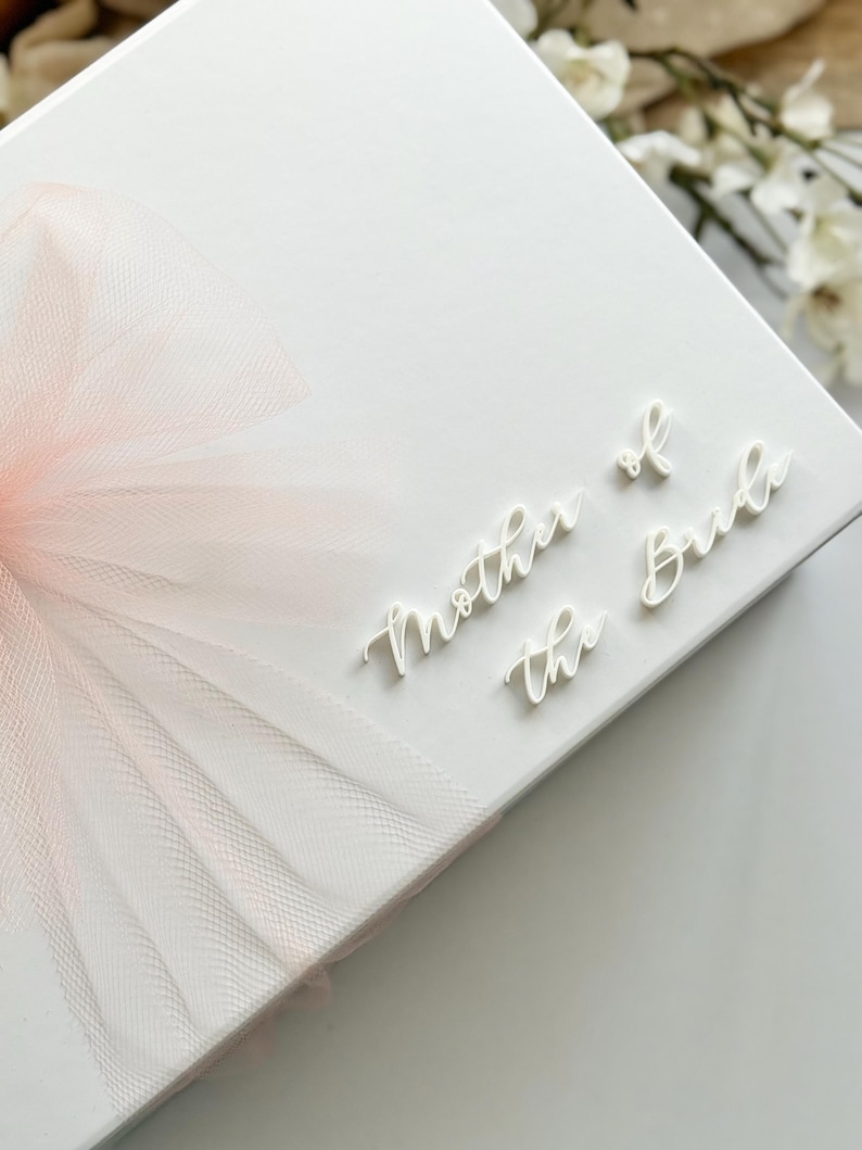 K&ouml;nnte beinhalten: Eine wei&szlig;e Geschenkbox mit einer zarten rosa T&uuml;llschleife. Die Worte &bdquo;Mother of the Bride&ldquo; sind in Schreibschrift oben rechts geschrieben. Eine Hochzeitsgeschenkbox f&uuml;r einen besonderen Anlass.
