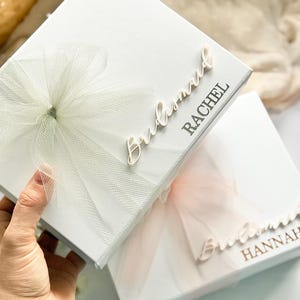 May include: Two white gift boxes, one held by a hand, with tulle bows and the word "Bridesmaid" in cursive. The first box has "Rachel" printed on it, and the second box has "Hannah" printed on it. The bows are white and light pink.