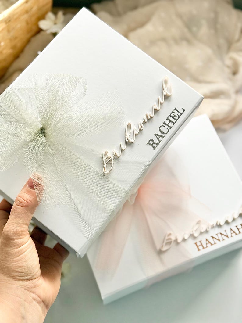 May include: Two white gift boxes, one held by a hand, with tulle bows and the word "Bridesmaid" in cursive. The first box has "Rachel" printed on it, and the second box has "Hannah" printed on it. The bows are white and light pink.
