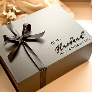 Könnte beinhalten: Eine dunkelgraue Geschenkbox, gebunden mit einem braunen Satinband und einer Schleife. Auf der Box steht in eleganter Schrift "TO MY Husband ON OUR WEDDING DAY". Die Box hat eine quadratische Form und ist wahrscheinlich für ein Hochzeitsgeschenk.