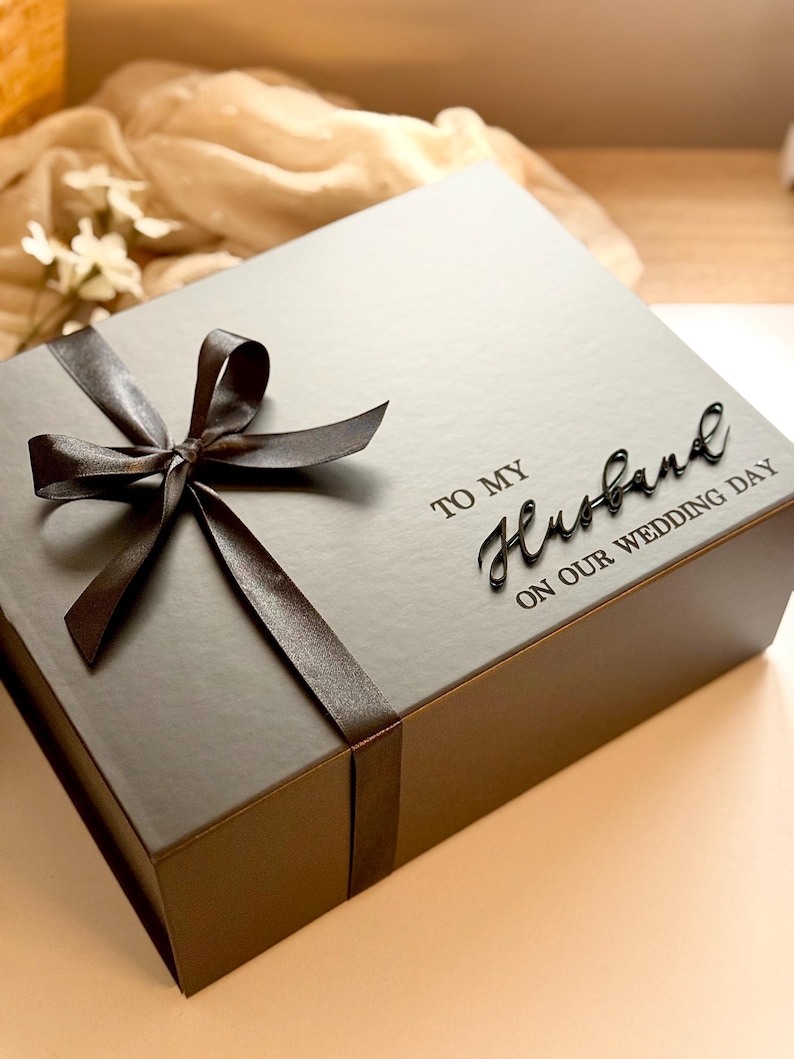 Puede incluir: Una caja de regalo gris oscuro atada con una cinta y un lazo de sat&eacute;n marr&oacute;n. La caja tiene las palabras "TO MY Husband ON OUR WEDDING DAY" en una elegante escritura. La caja es de forma cuadrada y parece estar hecha de cart&oacute;n.