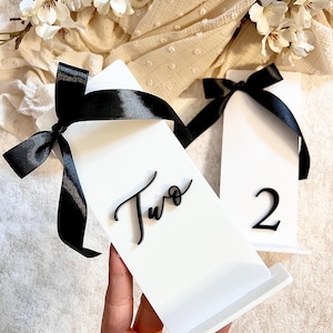 Könnte beinhalten: Zwei weiße Acryl-Tischnummernhalter mit schwarzen Schleifen. Ein Halter hat die Nummer "Zwei" in Kursivschrift und der andere die Nummer "2" in einer serifenlosen Schrift.