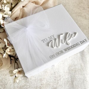May include: A white gift box with a tulle bow. The box has the words "TO MY Wife ON OUR WEDDING DAY" printed on the top. White flowers and a cream-colored cloth are in the background.