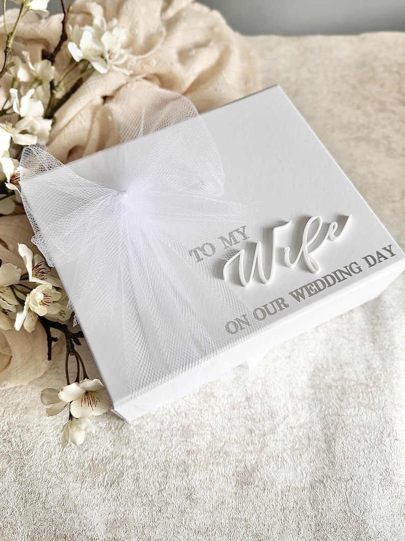 May include: A white gift box with a tulle bow. The box has the words "TO MY Wife ON OUR WEDDING DAY" printed on the top. White flowers and a cream-colored cloth are in the background.