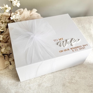 May include: A white gift box with a sheer white bow on top. The box is inscribed with the words "TO MY Wife ON OUR WEDDING DAY 5.21.2026" in gold lettering. The box is likely for a wedding gift.