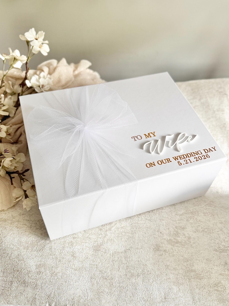 May include: A white gift box with a sheer white bow on top. The box is inscribed with the words "TO MY Wife ON OUR WEDDING DAY 5.21.2026" in gold lettering. The box is likely for a wedding gift.