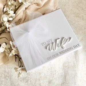 May include: A white square wedding card with the words "TO MY WIFE ON OUR WEDDING DAY" in elegant script. The card is adorned with a delicate white tulle bow. White flowers and soft fabric are in the background.