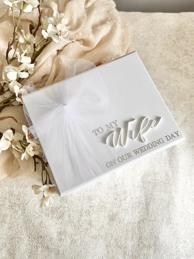 May include: A white square wedding card with the words "TO MY WIFE ON OUR WEDDING DAY" in elegant script. The card is adorned with a delicate white tulle bow. White flowers and soft fabric are in the background.