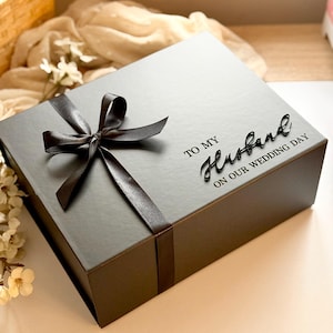 Puede incluir: Una caja de regalo gris oscuro atada con una cinta de sat&eacute;n negra. La caja tiene las palabras "TO MY Husband ON OUR WEDDING DAY" impresas en la parte superior. La caja est&aacute; sobre una superficie de color claro.