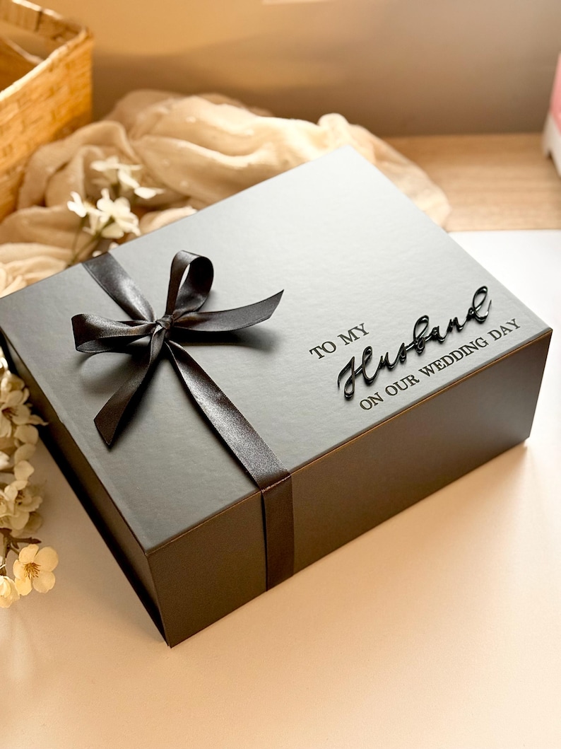 Puede incluir: Una caja de regalo gris oscuro atada con una cinta de sat&eacute;n negra. La caja tiene las palabras "TO MY Husband ON OUR WEDDING DAY" impresas en la parte superior. La caja est&aacute; sobre una superficie de color claro.