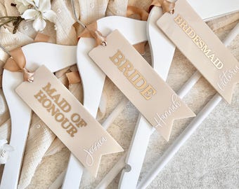 May include: White wooden hangers with beige ribbon and tags. The tags read "BRIDE", "BRIDESMAID", and "MAID OF HONOUR" in gold lettering, with names in cursive. Decorative flowers are in the background.