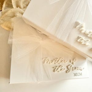 K&ouml;nnte beinhalten: Wei&szlig;e Geschenkbox mit dem Text "Mother of the Groom" und "MUM" in eleganter Schrift. Die Box ist mit einer wei&szlig;en Schleife und T&uuml;ll &uuml;berzogen, was auf ein Hochzeitsthema hindeutet.