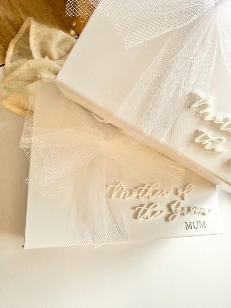 K&ouml;nnte beinhalten: Wei&szlig;e Geschenkbox mit dem Text "Mother of the Groom" und "MUM" in eleganter Schrift. Die Box ist mit einer wei&szlig;en Schleife und T&uuml;ll &uuml;berzogen, was auf ein Hochzeitsthema hindeutet.