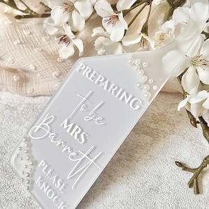 Puede incluir: Un colgador de puerta blanco esmerilado con una parte superior curva y un cuerpo rectangular. El texto "PREPARING to be MRS Barrett PLEASE KNOCK" se muestra en escritura blanca. Peque&ntilde;as perlas blancas est&aacute;n dispuestas a lo largo de los bordes.
