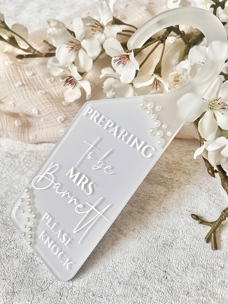 Puede incluir: Un colgador de puerta blanco esmerilado con una parte superior curva y un cuerpo rectangular. El texto "PREPARING to be MRS Barrett PLEASE KNOCK" se muestra en escritura blanca. Peque&ntilde;as perlas blancas est&aacute;n dispuestas a lo largo de los bordes.