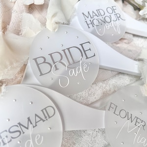 May include: White wooden hangers with round, clear acrylic tags. The tags have silver glitter and the text "Bride Sade", "Maid of Honour Cody", "Bridesmaid Jade", and "Flower Mia".