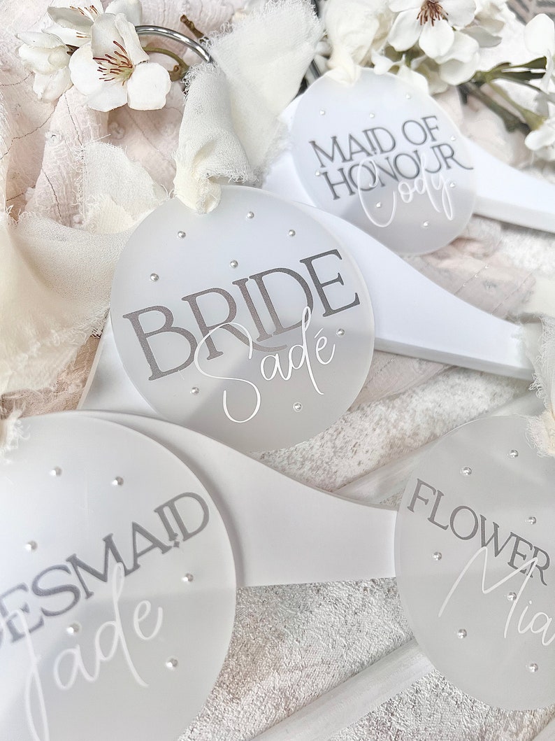 May include: White wooden hangers with round, clear acrylic tags. The tags have silver glitter and the text "Bride Sade", "Maid of Honour Cody", "Bridesmaid Jade", and "Flower Mia".