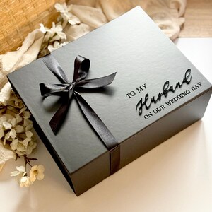 Puede incluir: Caja de regalo negra con una cinta de raso atada con un lazo. La caja tiene las palabras "TO MY Husband ON OUR WEDDING DAY" en una fuente de escritura. La caja est&aacute; sobre una superficie blanca con flores.
