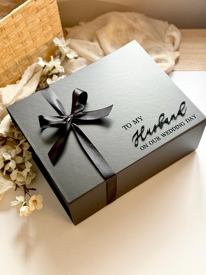 Puede incluir: Caja de regalo negra con una cinta de raso atada con un lazo. La caja tiene las palabras "TO MY Husband ON OUR WEDDING DAY" en una fuente de escritura. La caja est&aacute; sobre una superficie blanca con flores.