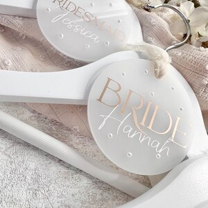 May include: Two white wooden hangers with round, clear acrylic tags. The top tag says "BRIDESMAID Jessica" and the bottom tag says "BRIDE Hannah".