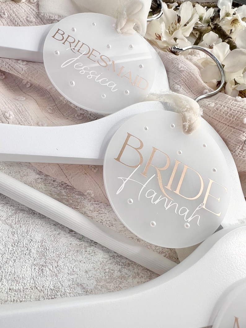May include: Two white wooden hangers with round, clear acrylic tags. The top tag says "BRIDESMAID Jessica" and the bottom tag says "BRIDE Hannah".