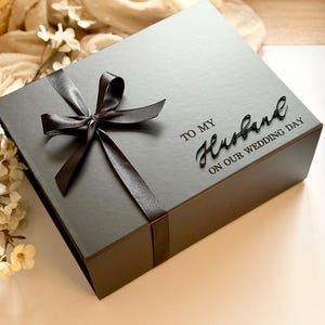 Puede incluir: Una caja de regalo gris oscuro atada con un lazo de cinta de sat&eacute;n. La caja est&aacute; inscrita con las palabras "TO MY Husband ON OUR WEDDING DAY" en una elegante escritura. Un regalo tem&aacute;tico de boda.