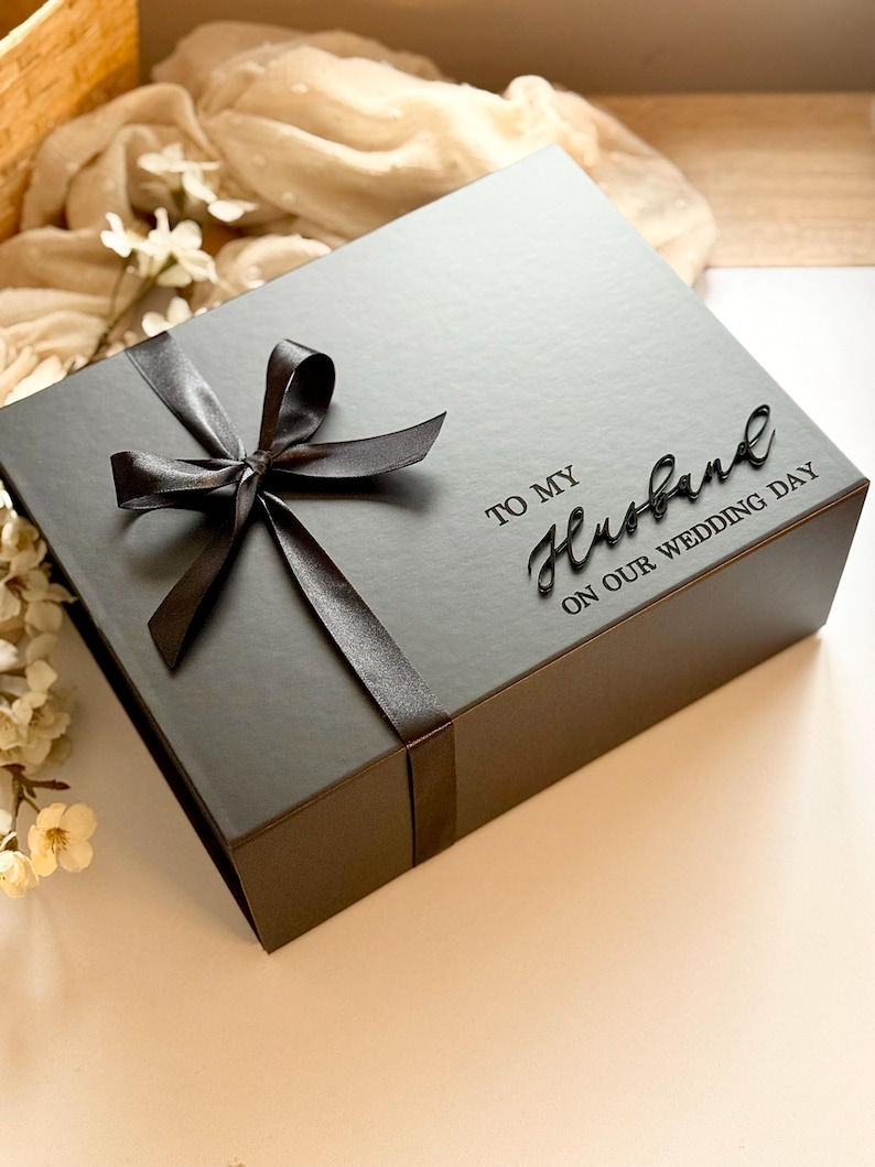 Puede incluir: Una caja de regalo gris oscuro atada con un lazo de cinta de sat&eacute;n. La caja est&aacute; inscrita con las palabras "TO MY Husband ON OUR WEDDING DAY" en una elegante escritura. Un regalo tem&aacute;tico de boda.