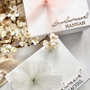 May include: Two white gift boxes with tulle bows and floral accents. One box has a light pink bow and the name "Hannah" in gold lettering. The other box has a white bow and the name "Rachel" in silver lettering. The boxes are for a bridal party.