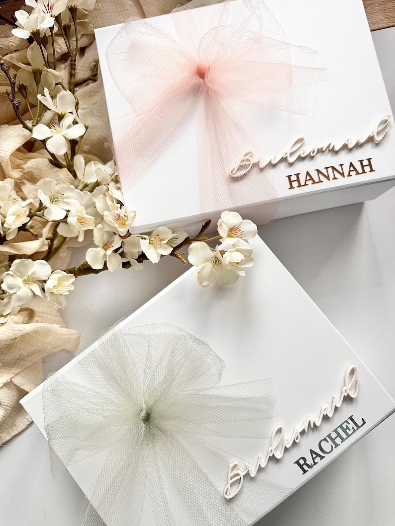 May include: Two white gift boxes with tulle bows and floral accents. One box has a light pink bow and the name "Hannah" in gold lettering. The other box has a white bow and the name "Rachel" in silver lettering. The boxes are for a bridal party.