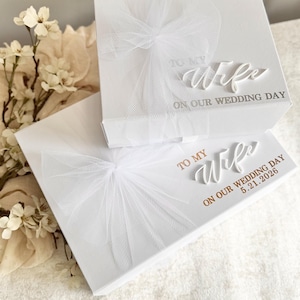 May include: Two white gift boxes with tulle bows. The top box reads "TO MY Wife ON OUR WEDDING DAY." The bottom box reads "TO MY Wife ON OUR WEDDING DAY 5.21.2026." The boxes are on a white surface.