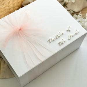 K&ouml;nnte beinhalten: Eine wei&szlig;e Geschenkbox mit einer zarten rosa T&uuml;llschleife. Die Worte "Mother of the Bride" sind in einer Schreibschrift auf der Box abgebildet. Die Box steht auf einer wei&szlig;en Oberfl&auml;che.