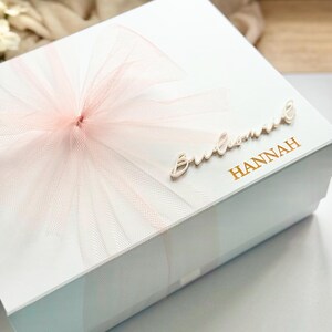 May include: A white gift box with a light pink tulle bow on top. The box has the word "Bridesmaid" in cursive and the name "Hannah" in gold lettering. The box is likely for a wedding or bridal party.