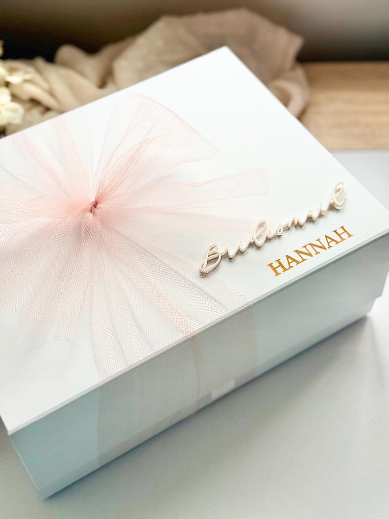 May include: A white gift box with a light pink tulle bow on top. The box has the word "Bridesmaid" in cursive and the name "Hannah" in gold lettering. The box is likely for a wedding or bridal party.