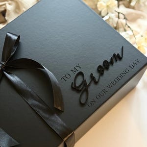May include: A black gift box tied with a black satin ribbon. The box has the words "TO MY Groom ON OUR WEDDING DAY" in a script font.