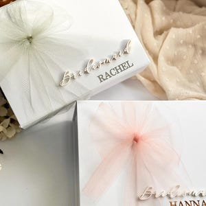 May include: Two white gift boxes with tulle bows. One box has a white bow and the name "Rachel" printed on it. The other box has a pink bow and the name "Hanna" printed on it. The word "Bridesmaid" is printed on both boxes.