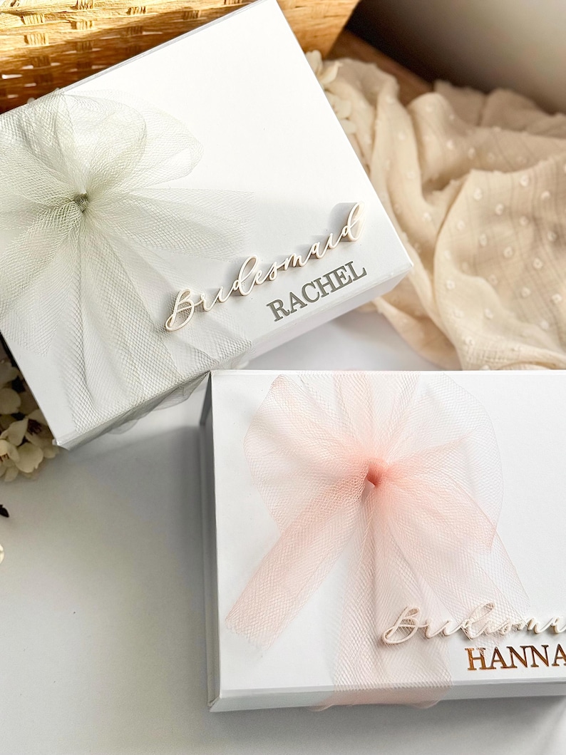 May include: Two white gift boxes with tulle bows. One box has a white bow and the name "Rachel" printed on it. The other box has a pink bow and the name "Hanna" printed on it. The word "Bridesmaid" is printed on both boxes.