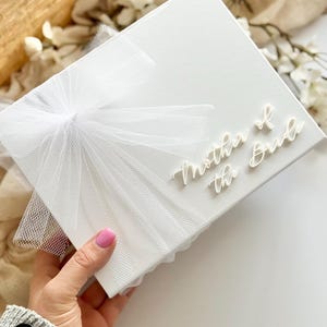 K&ouml;nnte beinhalten: Eine wei&szlig;e Geschenkbox mit einer T&uuml;llschleife und dem Schriftzug "Mother of the Bride" in zarter Schrift. Die Box wird von einer Person gehalten. Ein Geschenk zum Thema Hochzeit.