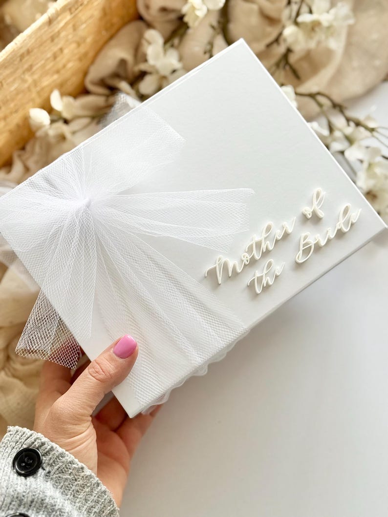 K&ouml;nnte beinhalten: Eine wei&szlig;e Geschenkbox mit einer T&uuml;llschleife und dem Schriftzug "Mother of the Bride" in zarter Schrift. Die Box wird von einer Person gehalten. Ein Geschenk zum Thema Hochzeit.
