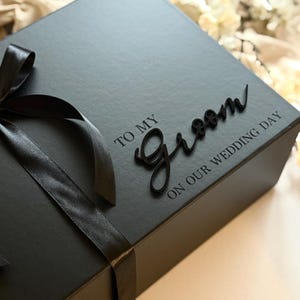 May include: A black gift box tied with a black satin ribbon and bow. The box has the words "TO MY Groom ON OUR WEDDING DAY" in a script font. The box is likely for a wedding.