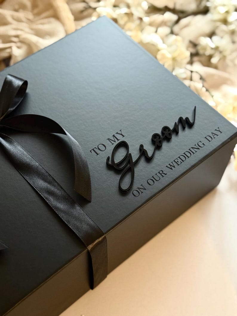 May include: A black gift box tied with a black satin ribbon and bow. The box has the words "TO MY Groom ON OUR WEDDING DAY" in a script font. The box is likely for a wedding.
