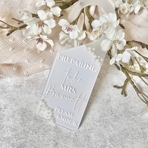 Puede incluir: Un colgador de puerta rectangular blanco con la inscripci&oacute;n "Preparing to be Mrs. Barrett, Please Knock". El colgador est&aacute; adornado con peque&ntilde;as perlas blancas y rodeado de flores blancas.
