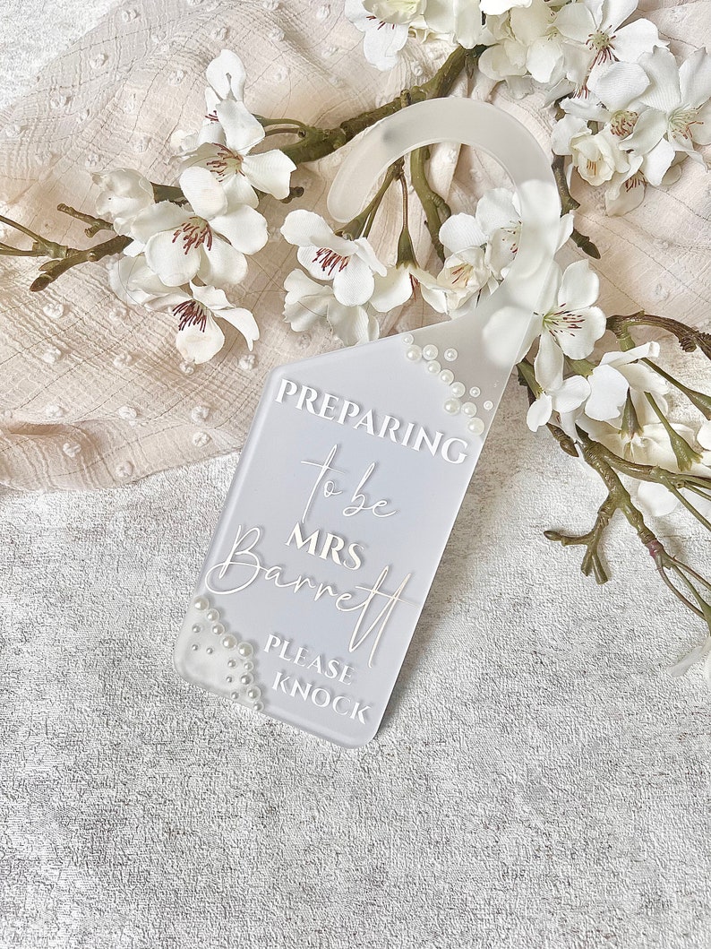 Puede incluir: Un colgador de puerta rectangular blanco con la inscripci&oacute;n "Preparing to be Mrs. Barrett, Please Knock". El colgador est&aacute; adornado con peque&ntilde;as perlas blancas y rodeado de flores blancas.