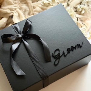 May include: A black gift box tied with a black satin ribbon and bow. The word "Groom" is written in a cursive font on the box. The box is on a white surface.
