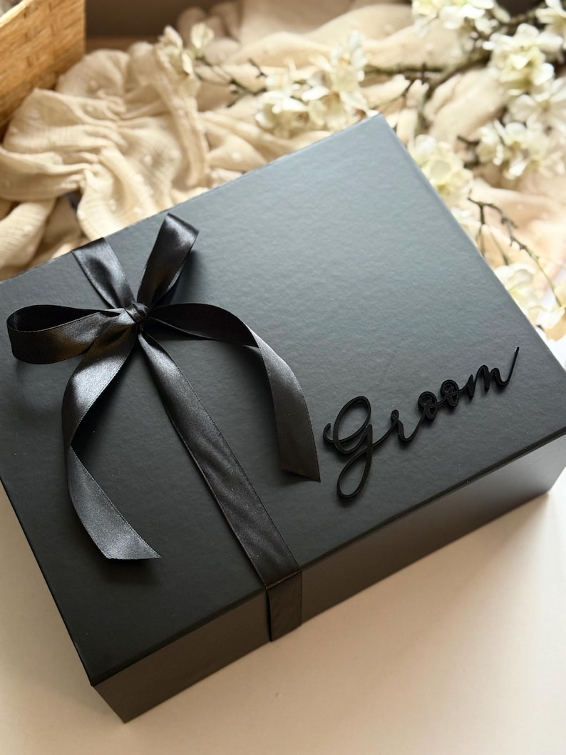 May include: A black gift box tied with a black satin ribbon and bow. The word "Groom" is written in a cursive font on the box. The box is on a white surface.