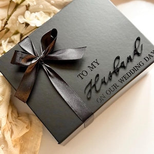 Puede incluir: Una caja de regalo negra con una cinta y un lazo de sat&eacute;n. La caja est&aacute; inscrita con las palabras "TO MY Husband ON OUR WEDDING DAY". La caja est&aacute; sobre una superficie blanca con flores.