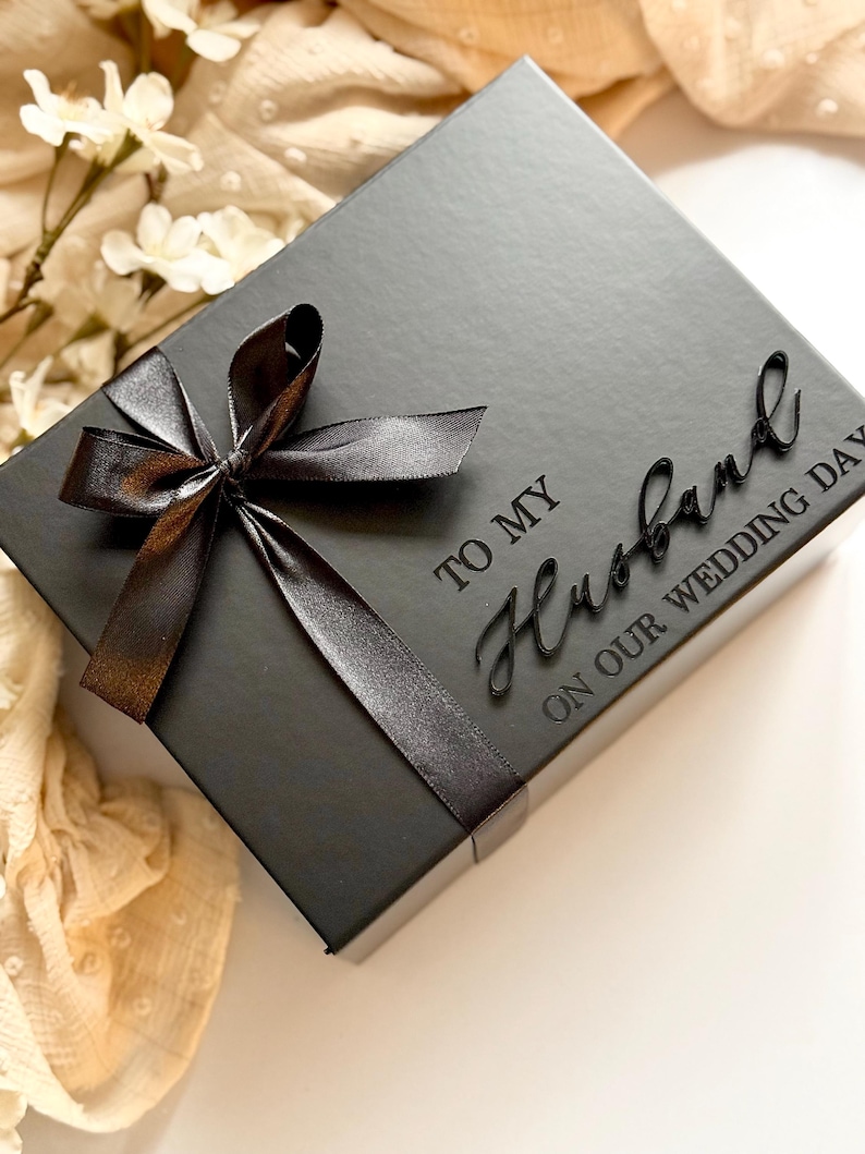 Puede incluir: Una caja de regalo negra con una cinta y un lazo de sat&eacute;n. La caja est&aacute; inscrita con las palabras "TO MY Husband ON OUR WEDDING DAY". La caja est&aacute; sobre una superficie blanca con flores.
